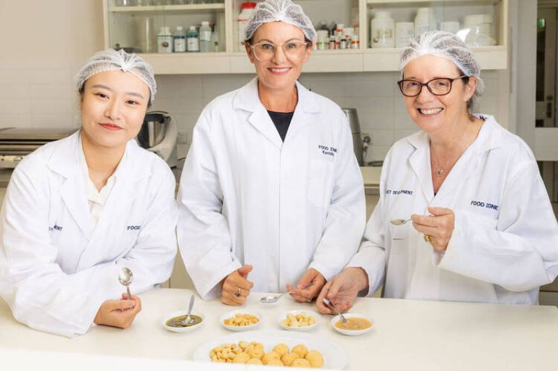 Three women wearing lab coats and hair nets stand behind a lab bench, which has macadamia samples on white plates