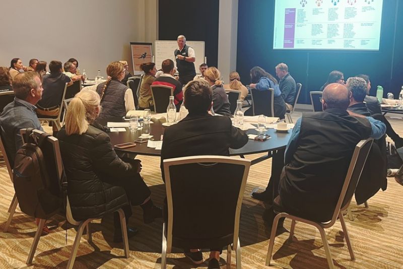 Workshop participants are seated around tables. In the background a presenter stands near a large screen with information on stakeholder review.
