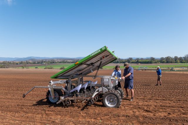 FarmDroid robot with raised solar panel in a broad, freshly worked field as two people review controls.