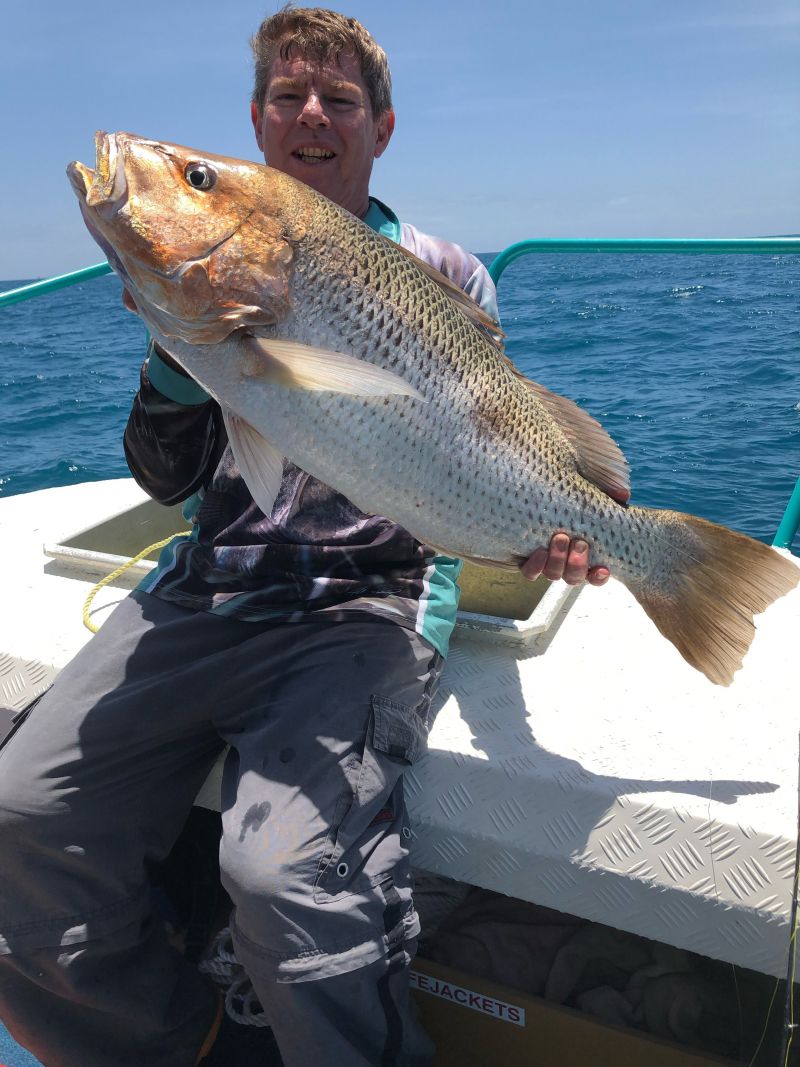 Garry Smith sits on a boat holding a large fish above the deck, with the ocean in the background.