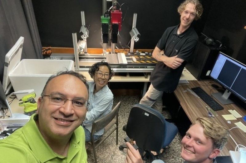 Four people smiling at the camera in a dark room with scientific equipment and a computer.