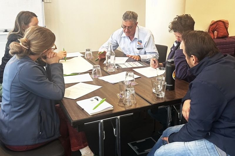 Workshop participants are gathered around a table listening to a facilitator whilst taking notes and reviewing papers. 
