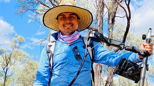 A man in a wide-brimmed hat holding field equipment in a rural Queensland landscape