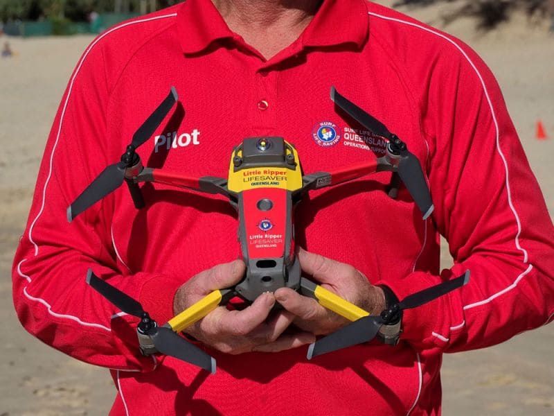 A man in a red shirt holding a yellow and red Little Ripper drone designed for SharkSmart trials on a beach.