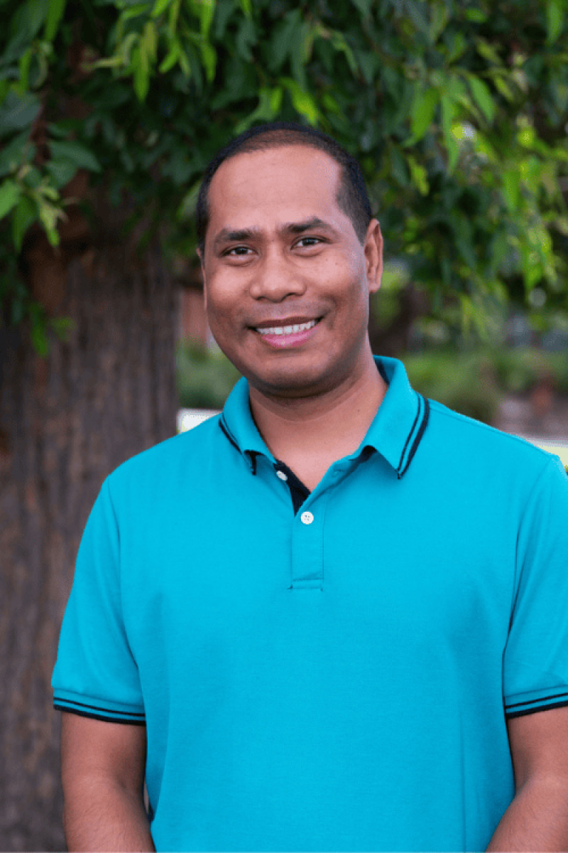 A man stands smiling, wearing a vibrant collared shirt with tree foliage in the background.