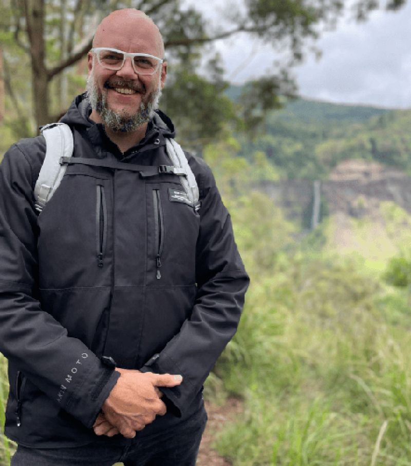Portrait of a Neil Davidson outdoors wearing white framed glasses and a black jacket.