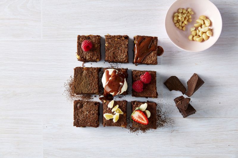 Brownies covered in chocolate, raspberries and strawberries with a bowl of macadamia nuts
