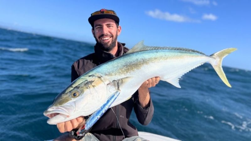 Man wearing a cap holds a large fish, smiling on a boat with choppy blue ocean waves under a clear sky.