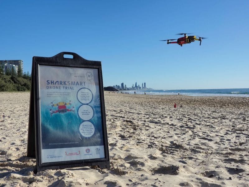 A SharkSmart Drone Trial sign on a beach with a Little Ripper drone flying nearby and the Gold Coast skyline in the distance.