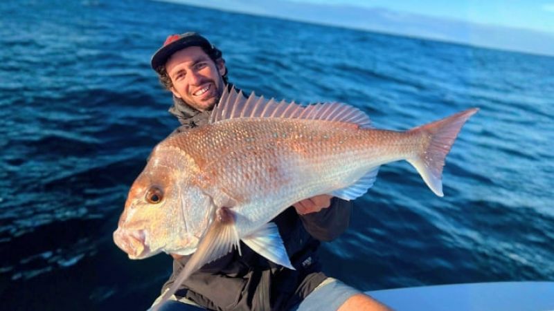 Man wearing a cap holds a large fish, smiling on a boat with choppy blue ocean waves under a clear sky.