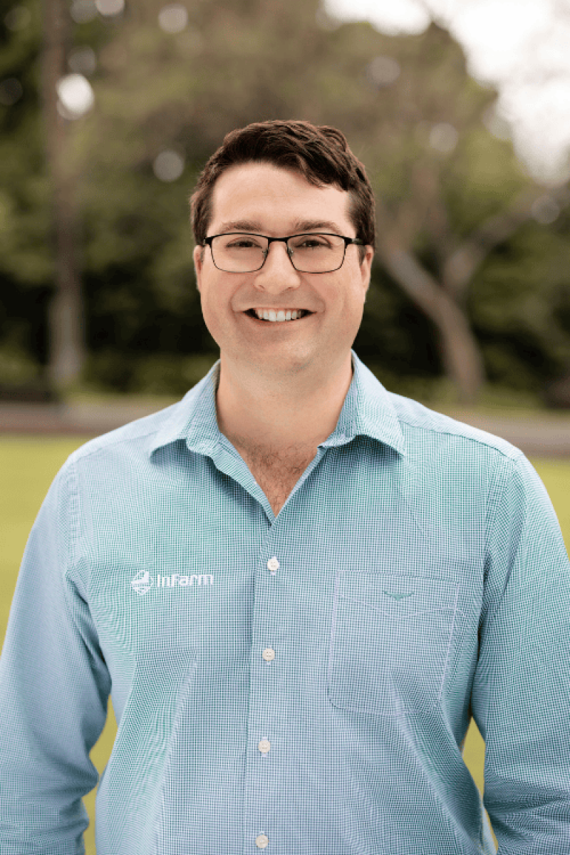 A man with dark hair stands in a grassy field with trees, wearing spectacles and a green button-up shirt with the word ‘InFarm’ on it. 