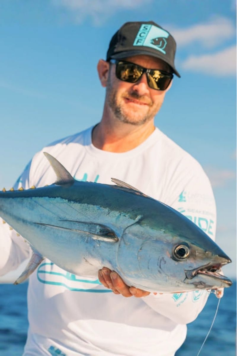 Man proudly holds a large tuna fish on a sunny day at sea. He wears a white shirt and black cap with sunglasses.