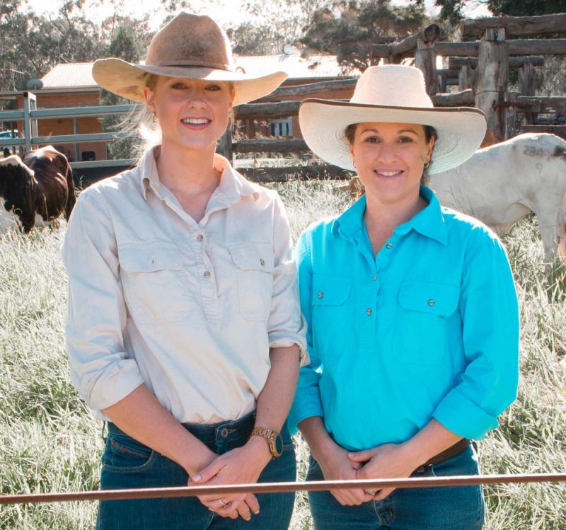 Two women stand smiling in a grassy field with cows in the background. The women wear wide-brimmed hats and collared work shirts.