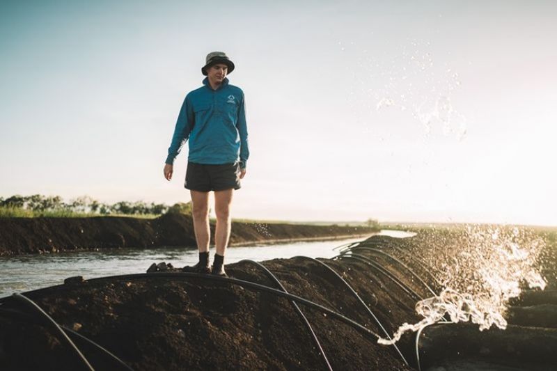 Farmer wearing blue shirt and a sunhat, standing on a dam wall. Irrigation hoses with water exiting 