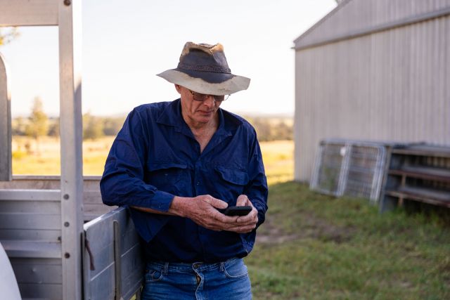 Older farmer in a hat leaning on a ute tray, looking at a smartphone beside a shed.