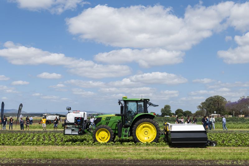 A green tractor with front and rear agricultural technology attachments operates in crop rows